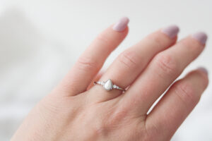 Bague en lait maternel en forme de goutte sertie de quatre pierres de naissance, tenue délicatement entre les doigts, mise en valeur par un fond neutre.