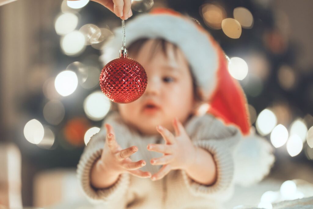 Bébé assis devant un sapin de Noël, portant un bonnet de père Noël et tentant d’attraper une boule de Noël.