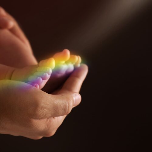 Pieds de bébé arc-en-ciel dans les mains d’un parent avec lumière colorée, symbole d’espoir, d’amour et de renaissance après une perte périnatale