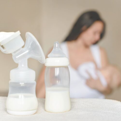 Tire-lait et biberon de lait maternel au premier plan, avec une mère allaitant son bébé en arrière-plan.