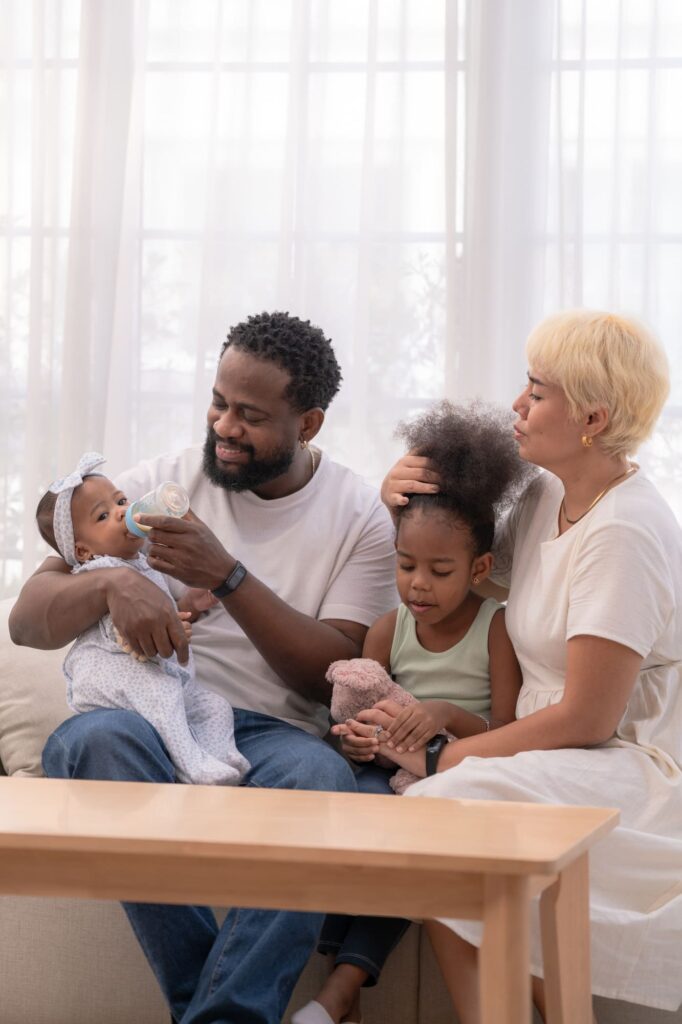Père donnant le biberon à son bébé, accompagné de sa conjointe et de leur jeune enfant dans le salon familial.